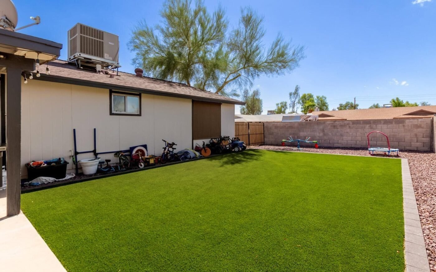 Image of a backyard in Tucson, AZ with a well-maintained artificial grass lawn. Two cornhole boards are placed on the grass, and a small dog is standing near one of the boards. The patio area is paved with light-colored stone tiles, and there are plants and rocks along the fence. Contact us for free quotes on artificial grass services.